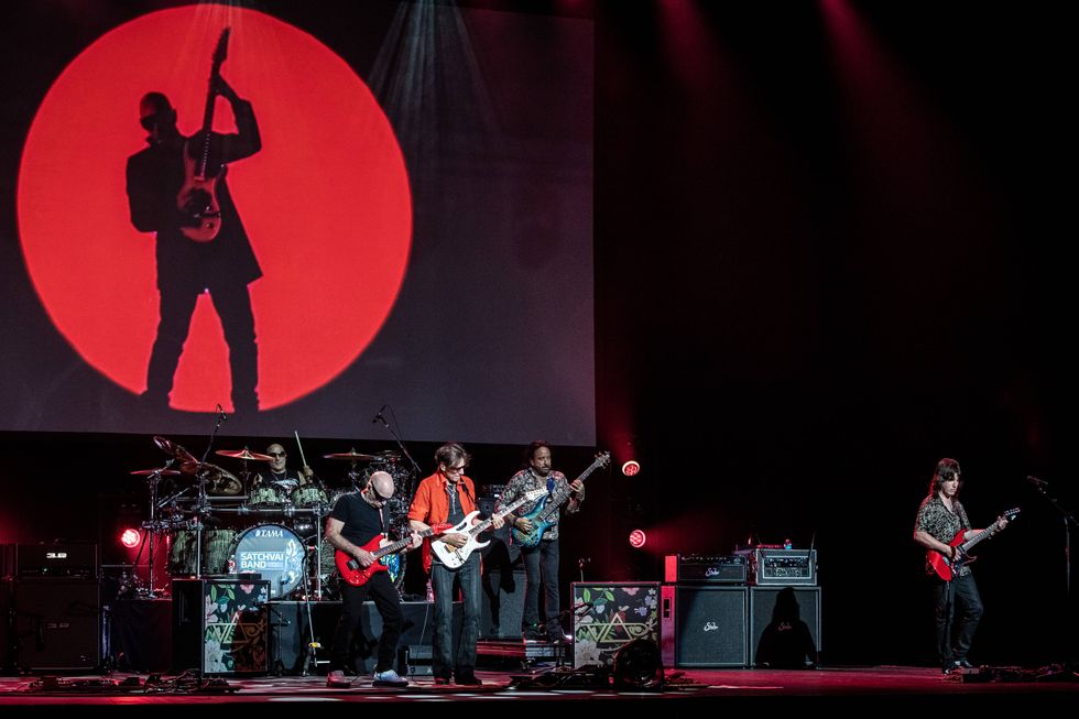 A rock band performs on stage with a large red backdrop and dramatic lighting.