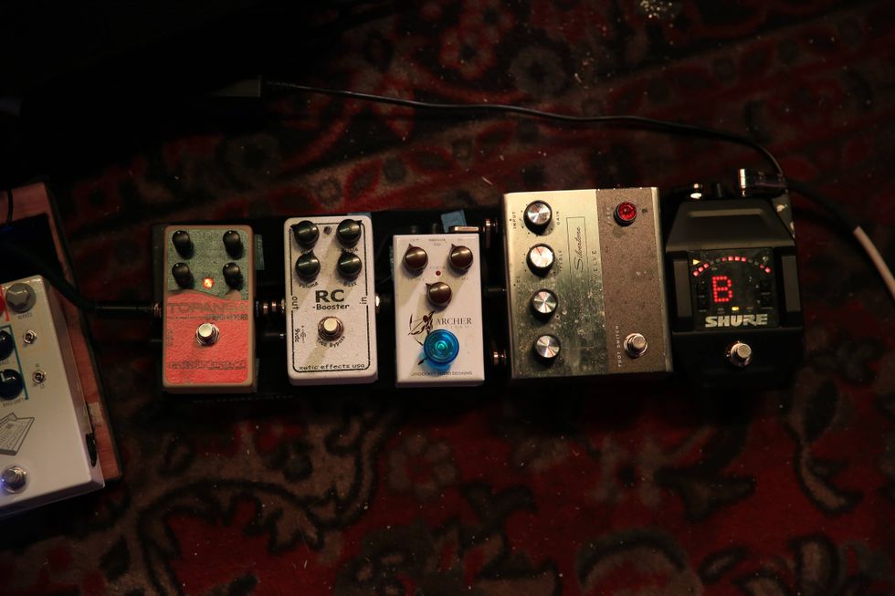 A row of guitar effect pedals on a patterned rug, with a tuner displaying "B."