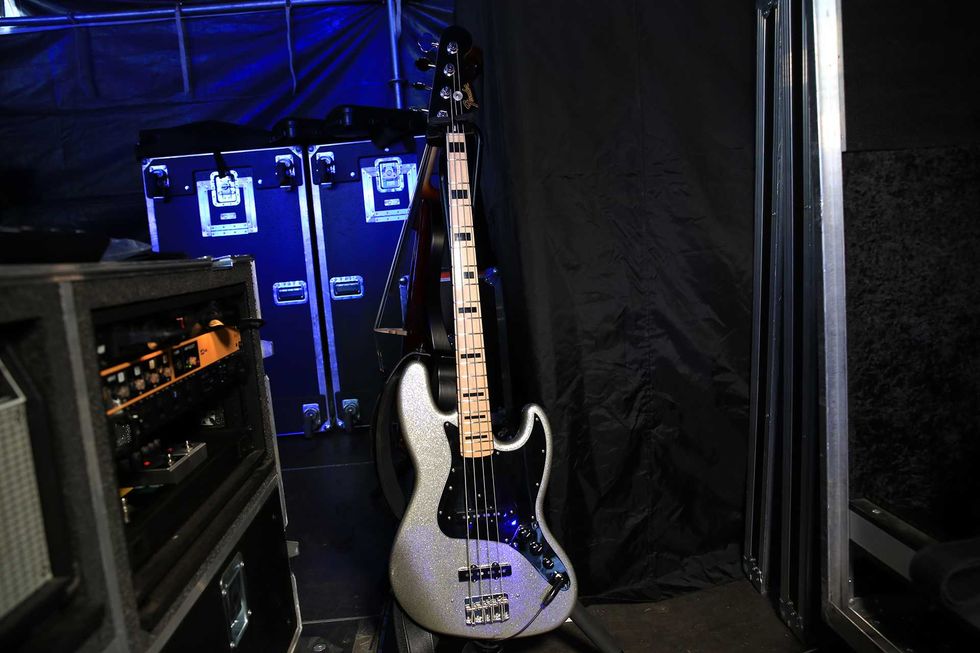 A shimmering silver bass guitar stands against a black backdrop with gear cases nearby.