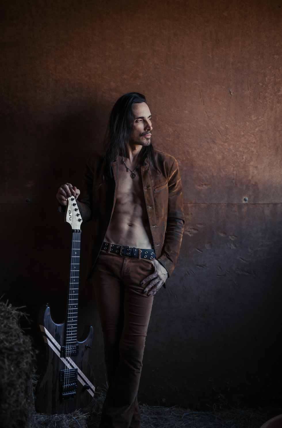 A shirtless man in a brown jacket leans against a wall, holding an electric guitar.
