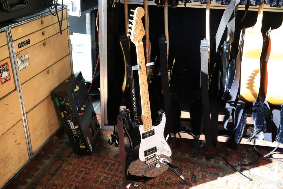 A silver electric guitar stands on a carpet, surrounded by other guitars and equipment.