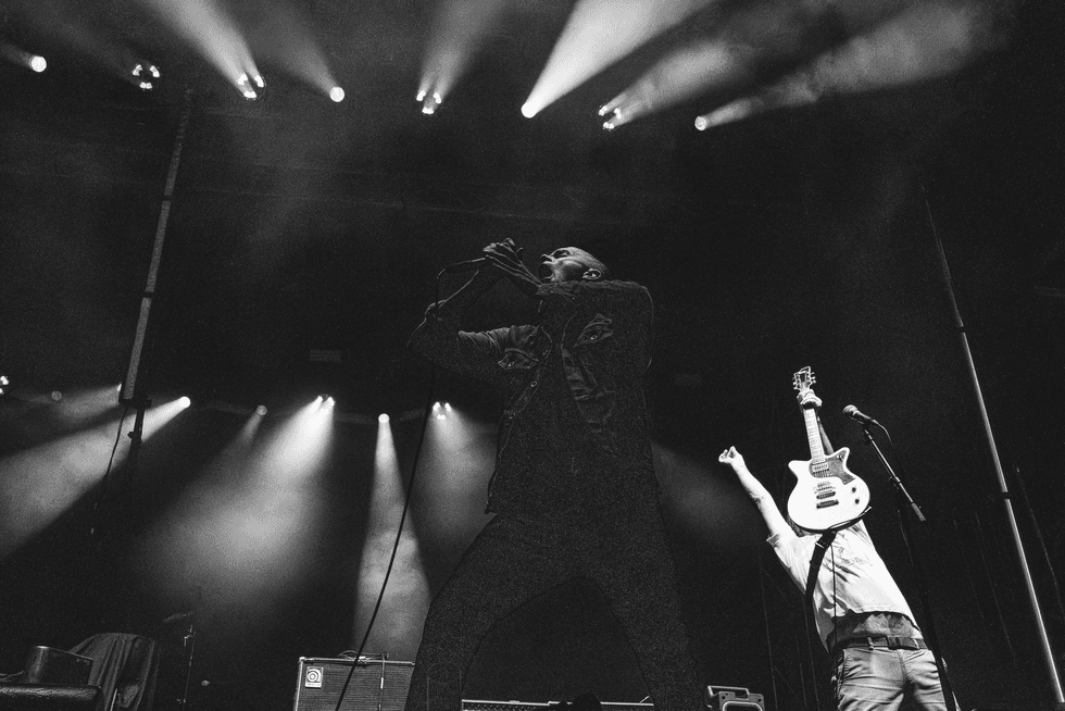 A singer passionately performs on stage while a guitarist raises his guitar, spotlights shining.