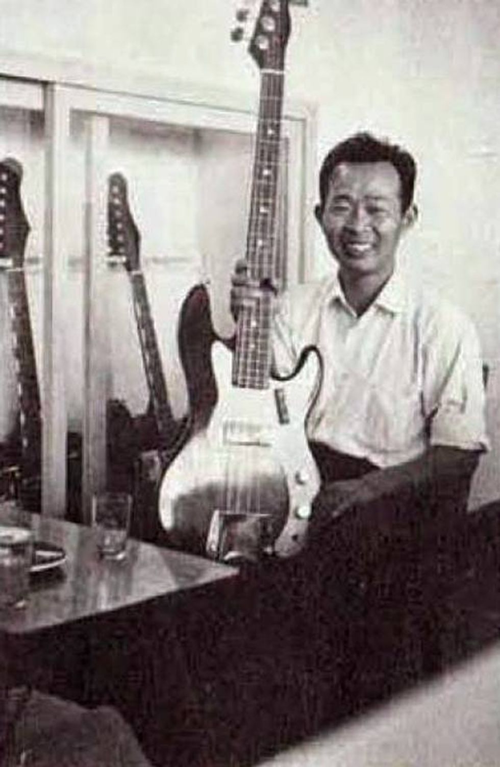 A smiling man holds a large guitar in a room with several guitars displayed.