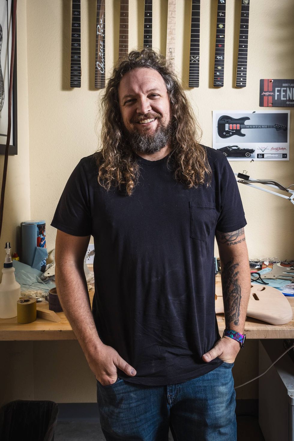 A smiling man with long hair stands in a workshop, surrounded by guitar parts and tools.