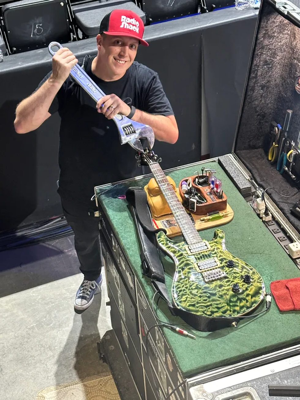 A smiling technician holds a large wrench beside a colorful guitar on a workbench.