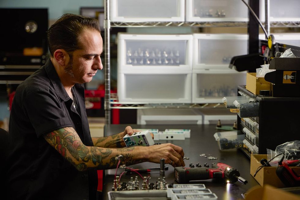 A tattooed man assembles equipment at a cluttered workbench in a workshop.