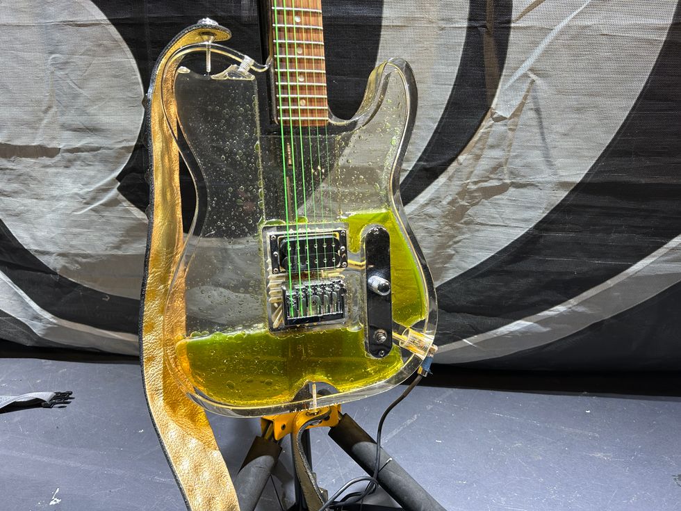 A transparent electric guitar with green strings and yellow liquid inside.