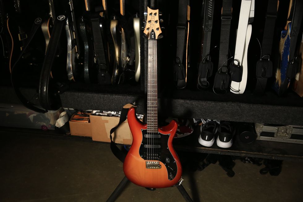 A vibrant orange electric guitar stands in front of a collection of other guitars.