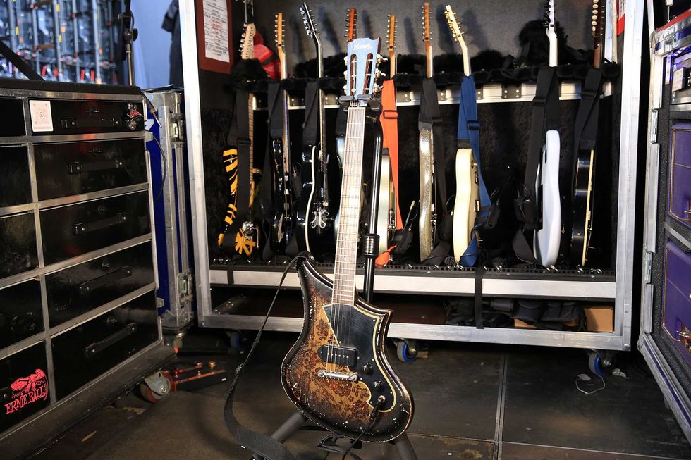 A vintage electric guitar stands in front of a case filled with various other guitars.