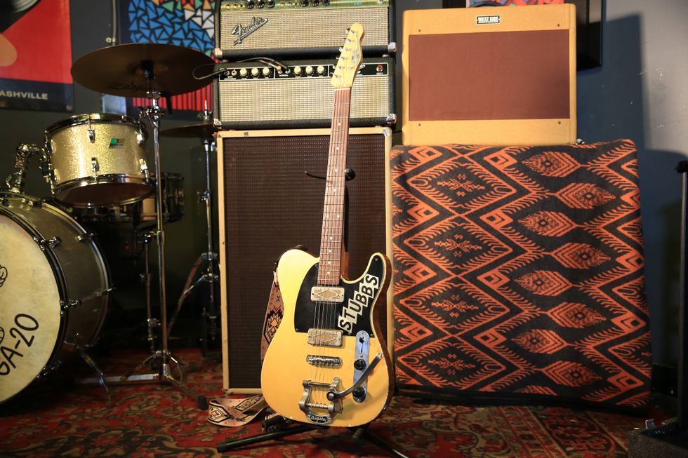 A vintage electric guitar with "STUBBS" sticker, next to drum set and amplifiers.