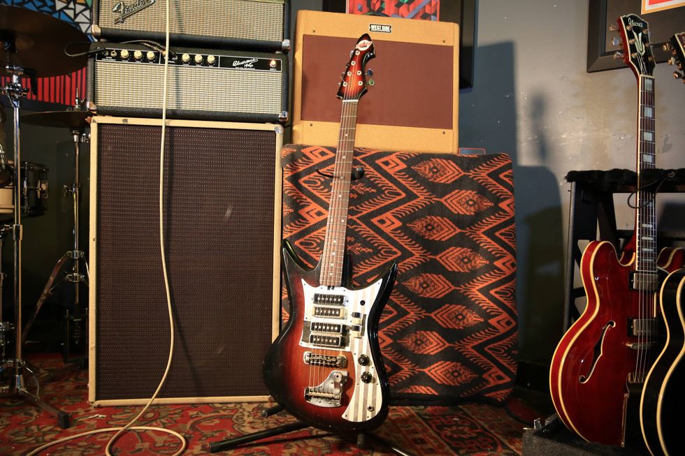 A vintage guitar stands beside amplifiers and drums in a music studio setting.