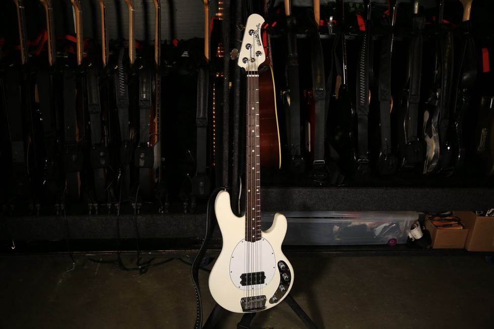 A white bass guitar stands in front of various other guitars on a rack.