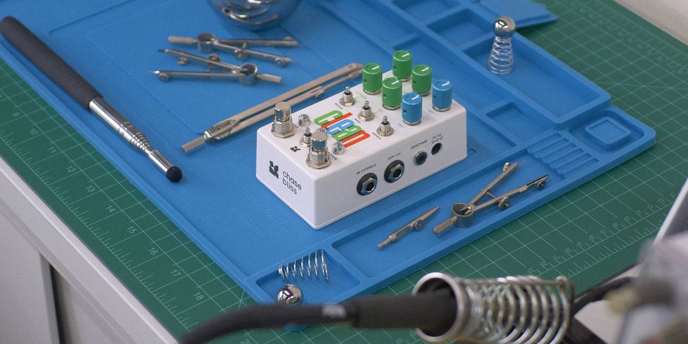 A white effects pedal surrounded by various tools on a blue work mat.