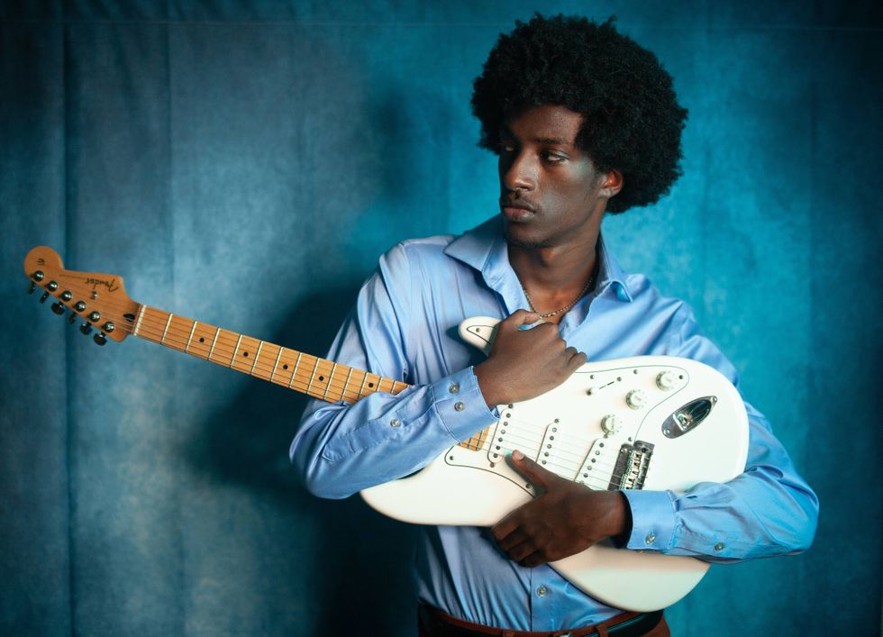 A young man with an afro poses with an electric guitar against a blue backdrop.