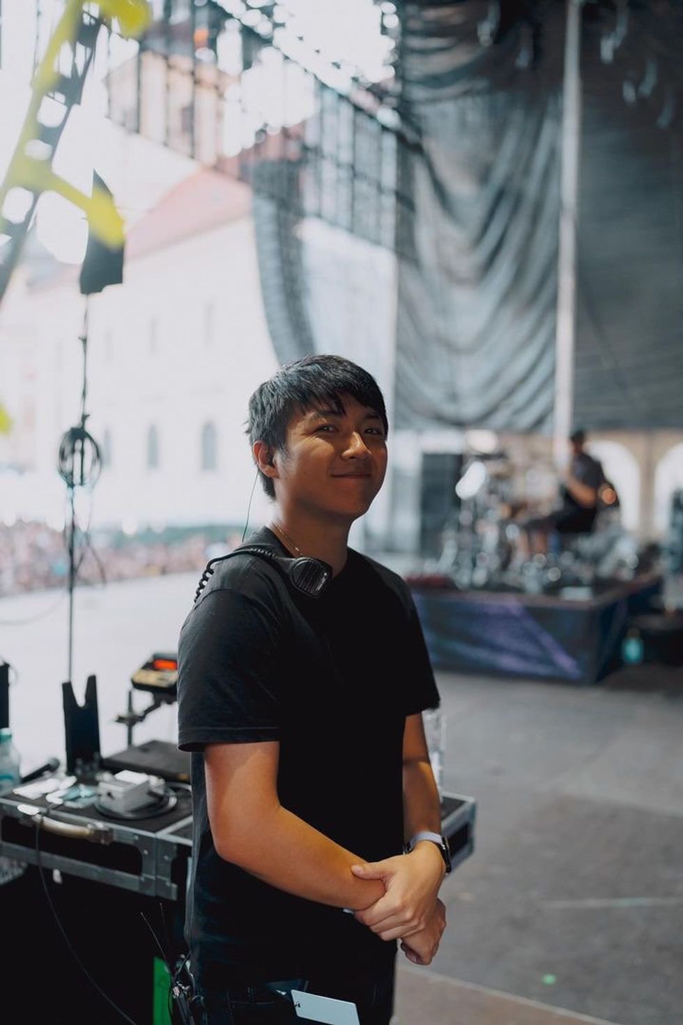 A young technician smiles backstage at a music festival, with equipment in the background.