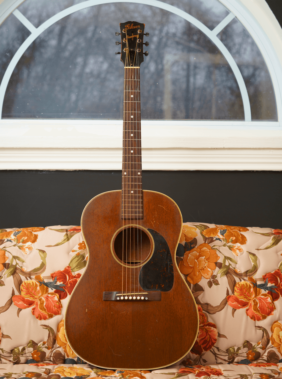 Acoustic guitar on a floral couch, with a large window in the background.