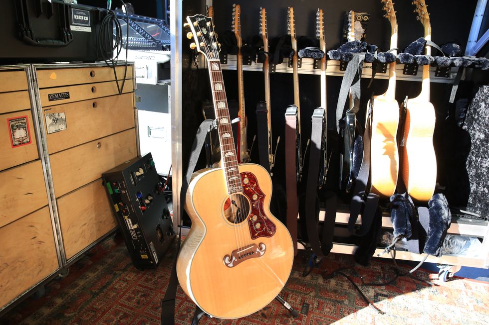 An acoustic guitar leans against a rack with multiple guitars and equipment in a backstage setting.