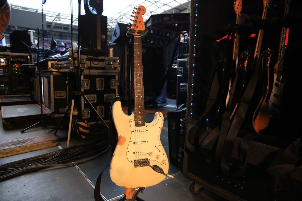 An aged white electric guitar stands on stage, surrounded by equipment and other guitars.