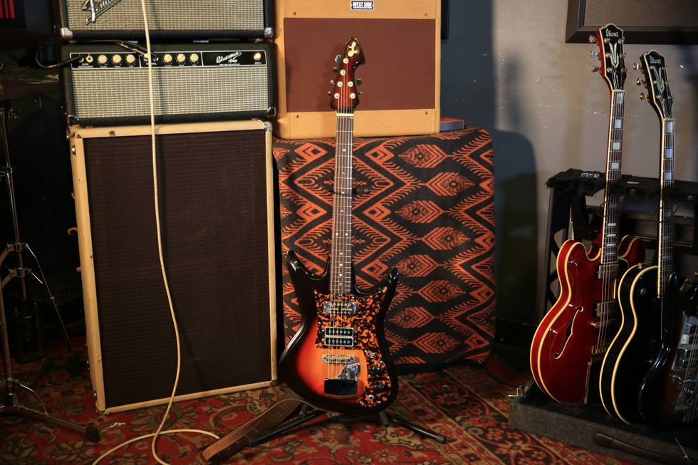 An electric guitar stands against amplifiers and other guitars in a music studio setting.