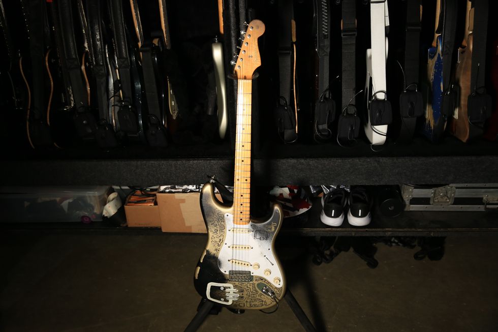 An electric guitar with intricate designs stands in front of a wall of other guitars.