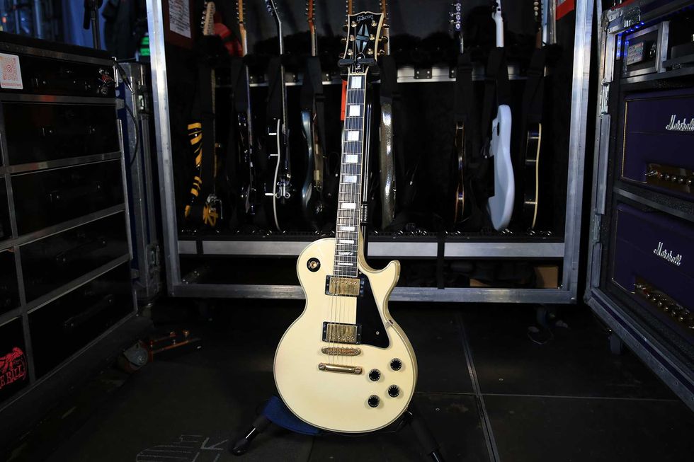 An off-white electric guitar stands in front of a backdrop of various other guitars.