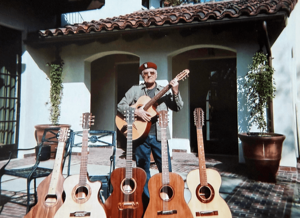 An older man in a beret plays guitar among several acoustic guitars on display.