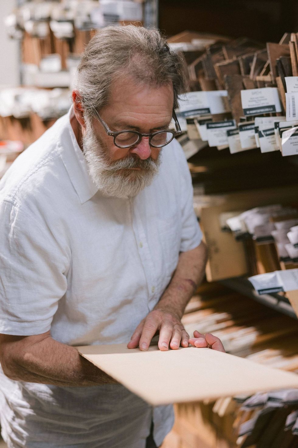 An older man with glasses examines a file in a cluttered archive or storage space.