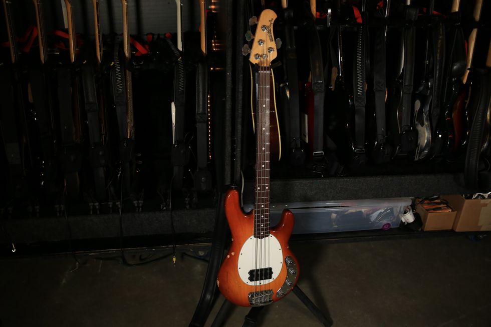 An orange bass guitar stands in front of a wall of other guitars, all in a dimly lit space.