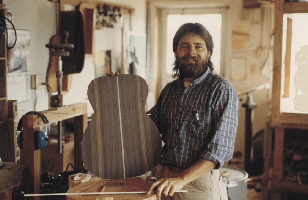 Bearded man in a workshop holds a guitar body, surrounded by tools and wood.