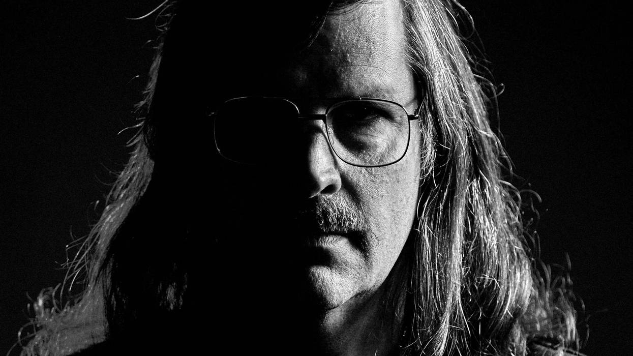 Black and white portrait of a man with long hair and glasses, shadowed and intense.