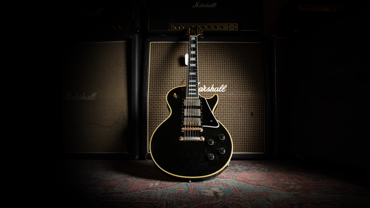Black electric guitar in front of two Marshall amplifiers on a patterned rug.