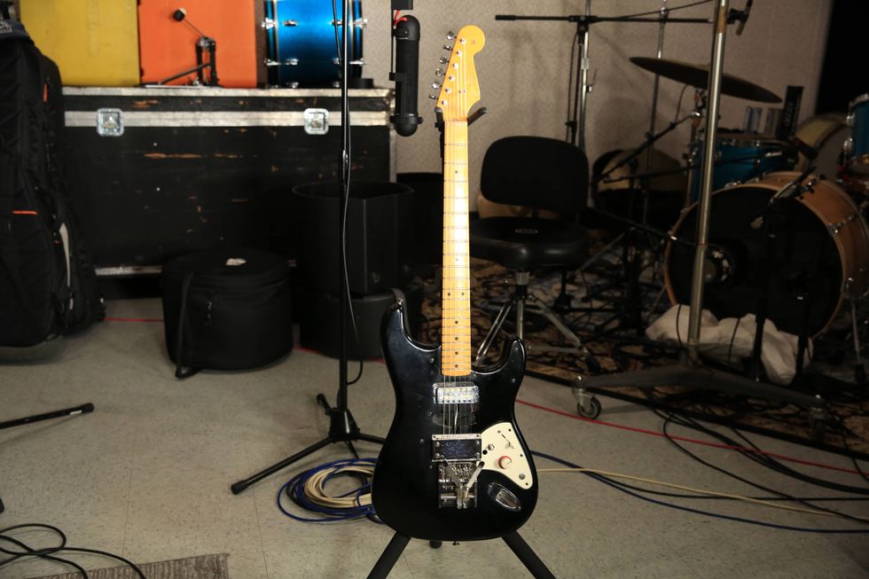 Black electric guitar on a stand in a music studio with drums in the background.
