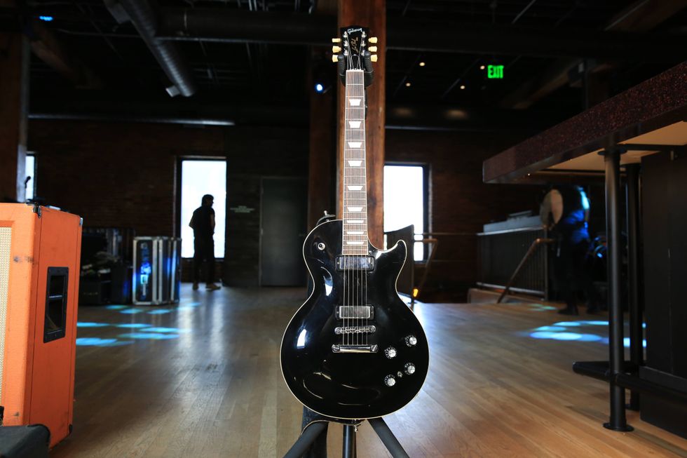 Black electric guitar on a stand, set in a dimly lit venue with wooden floors.