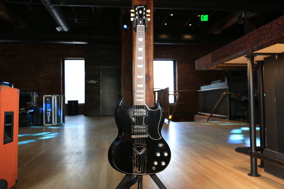 Black electric guitar stands on a stage in a well-lit wooden venue.