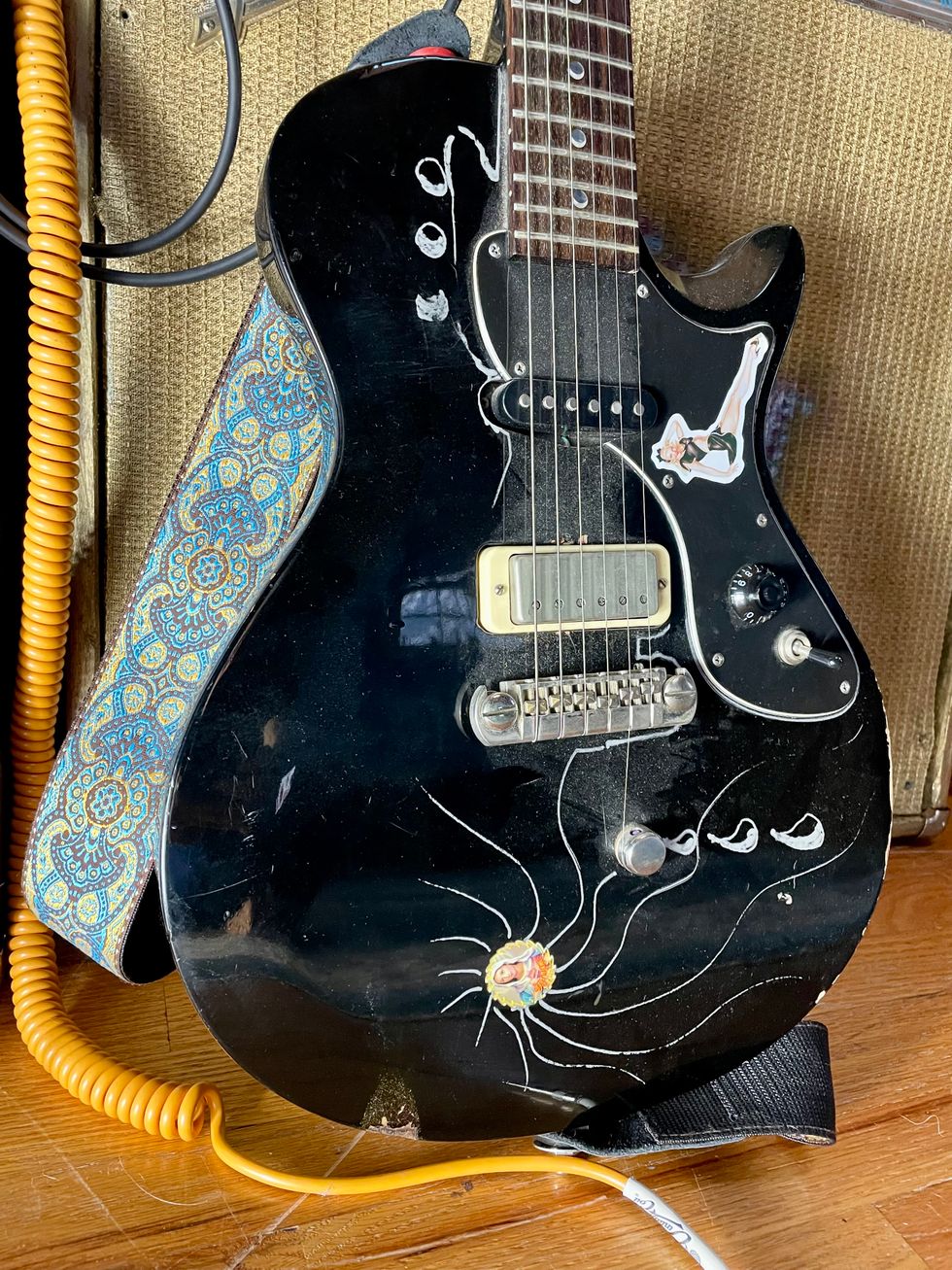 Black electric guitar with decorative stickers and a colorful strap, resting on an amp.