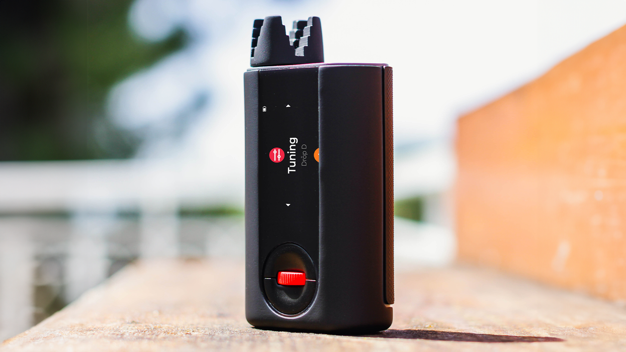 Black tuning device with a red button, displayed on a wooden surface against a blurred background.
