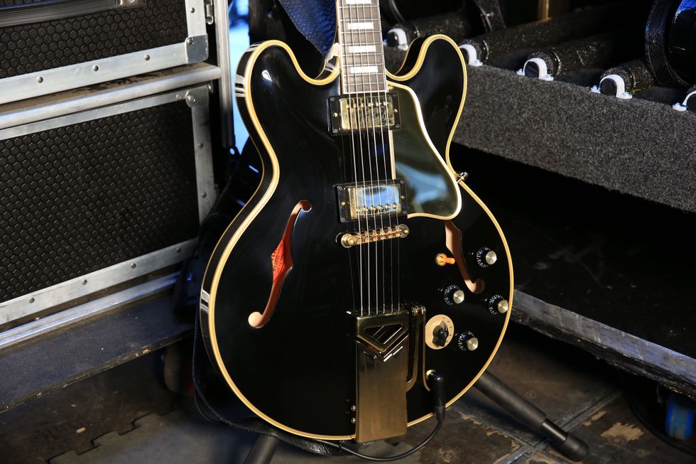 Close-up of a black electric guitar with gold accents, resting against a music stage backdrop.