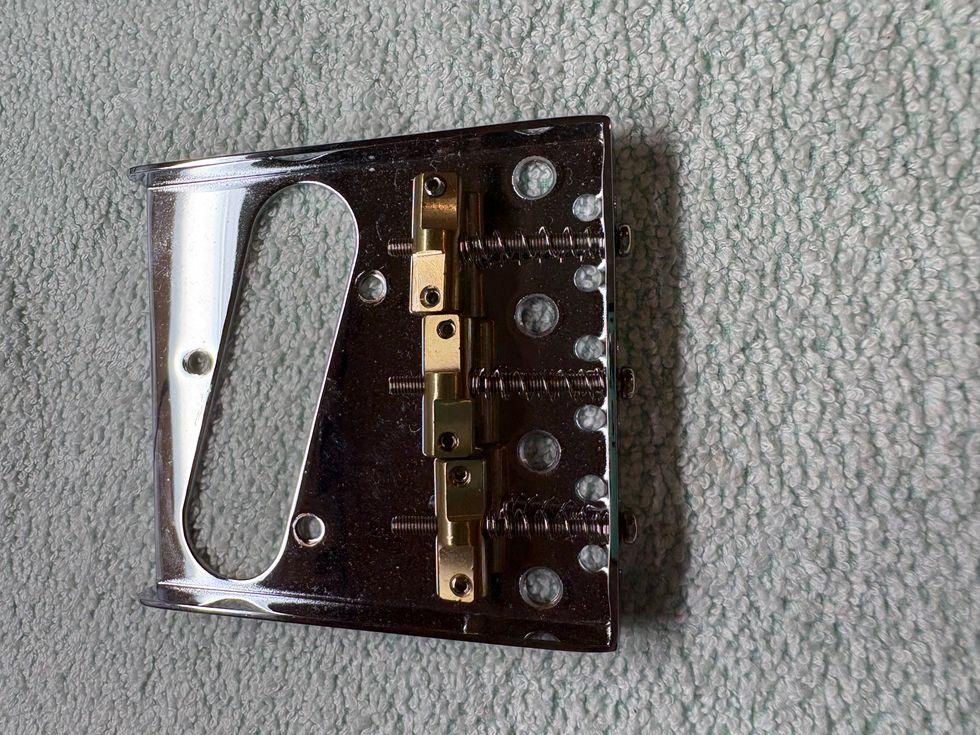 Close-up of a metallic guitar bridge with screws and mounting holes on a textured surface.