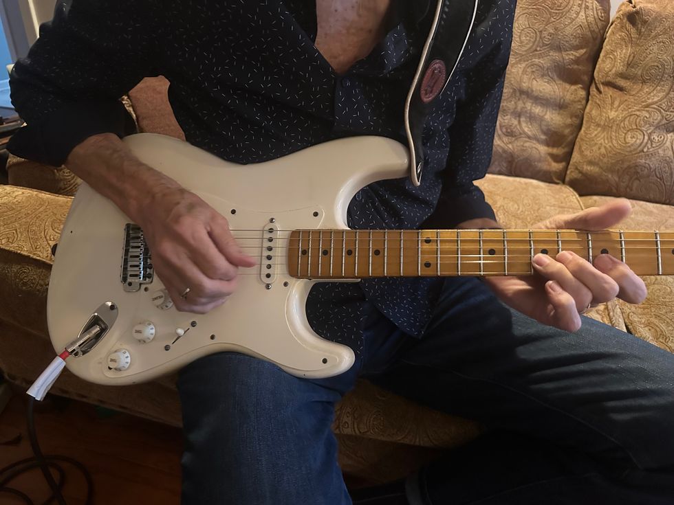 Close-up of a person playing a white electric guitar while seated on a couch.
