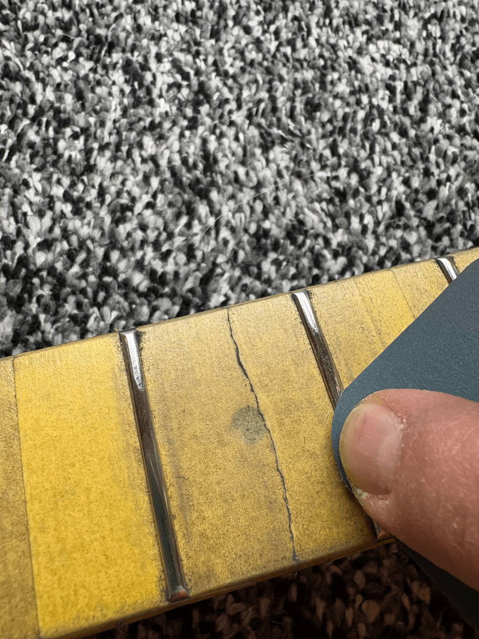 Close-up of a person's finger sanding a yellow guitar fretboard on gray carpet.