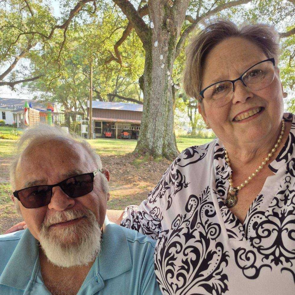 Elderly couple smiling together outdoors, surrounded by trees and a rural setting.