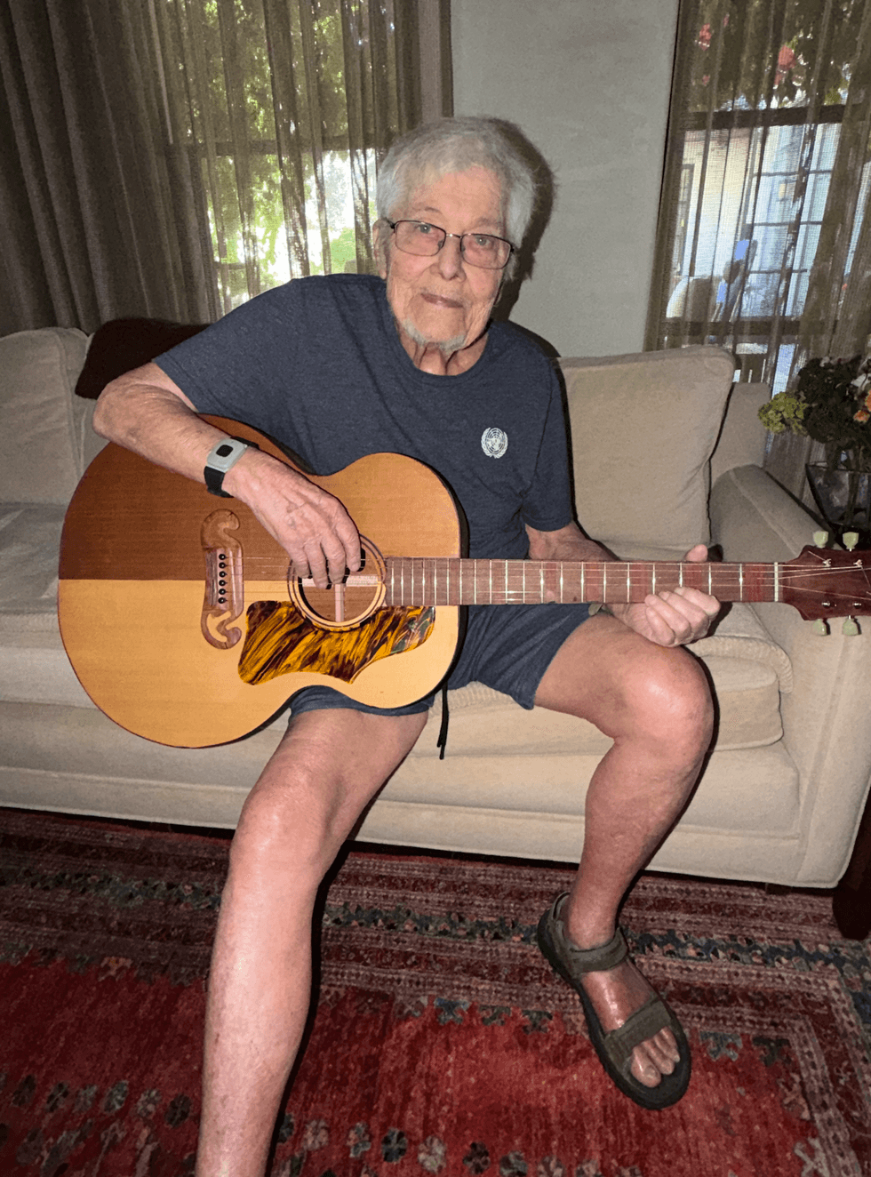 Elderly man wearing glasses plays an acoustic guitar while seated on a couch.