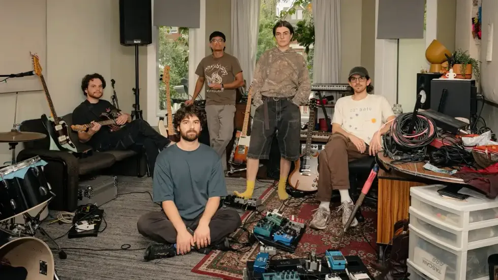 Five musicians in a cluttered room filled with instruments and equipment.