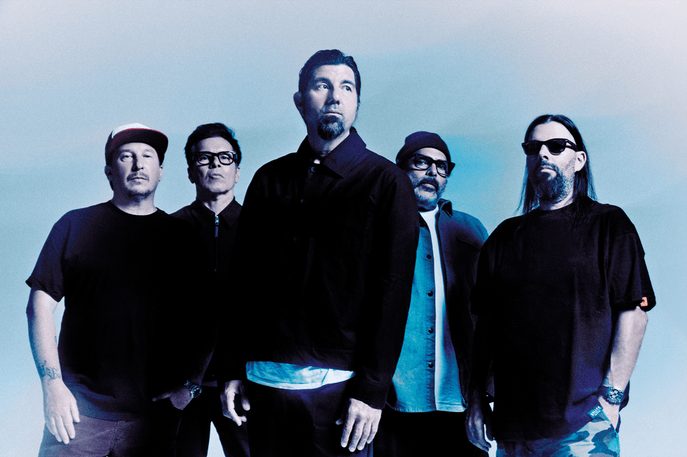 Five musicians stand together against a blue background, dressed in casual attire.