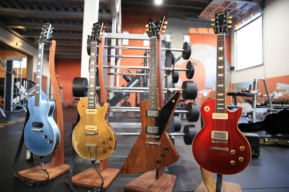 Four electric guitars displayed in a gym, with weights and exercise equipment in the background.