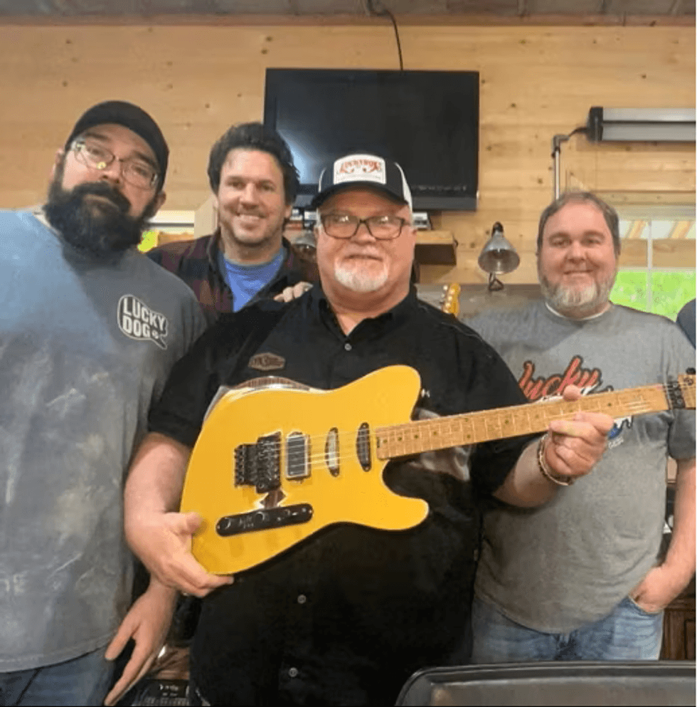 Four men in a workshop holding a yellow guitar, smiling together.