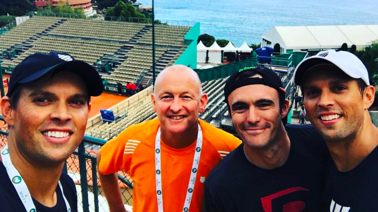 Four men smile for a selfie at a tennis venue overlooking the ocean.