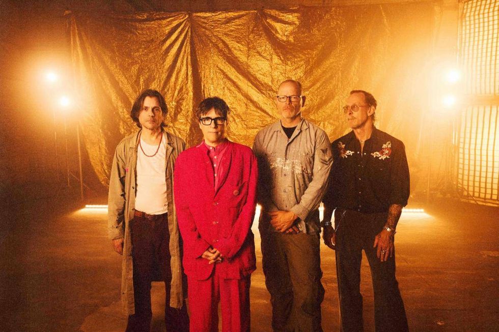 Four musicians pose in front of a golden backdrop, illuminated by warm stage lights.
