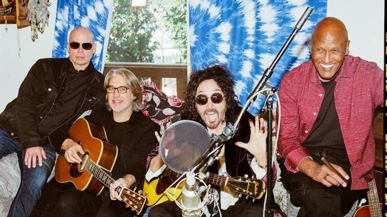 Four musicians sit together in a cozy room, playing guitars and smiling, with colorful decor.