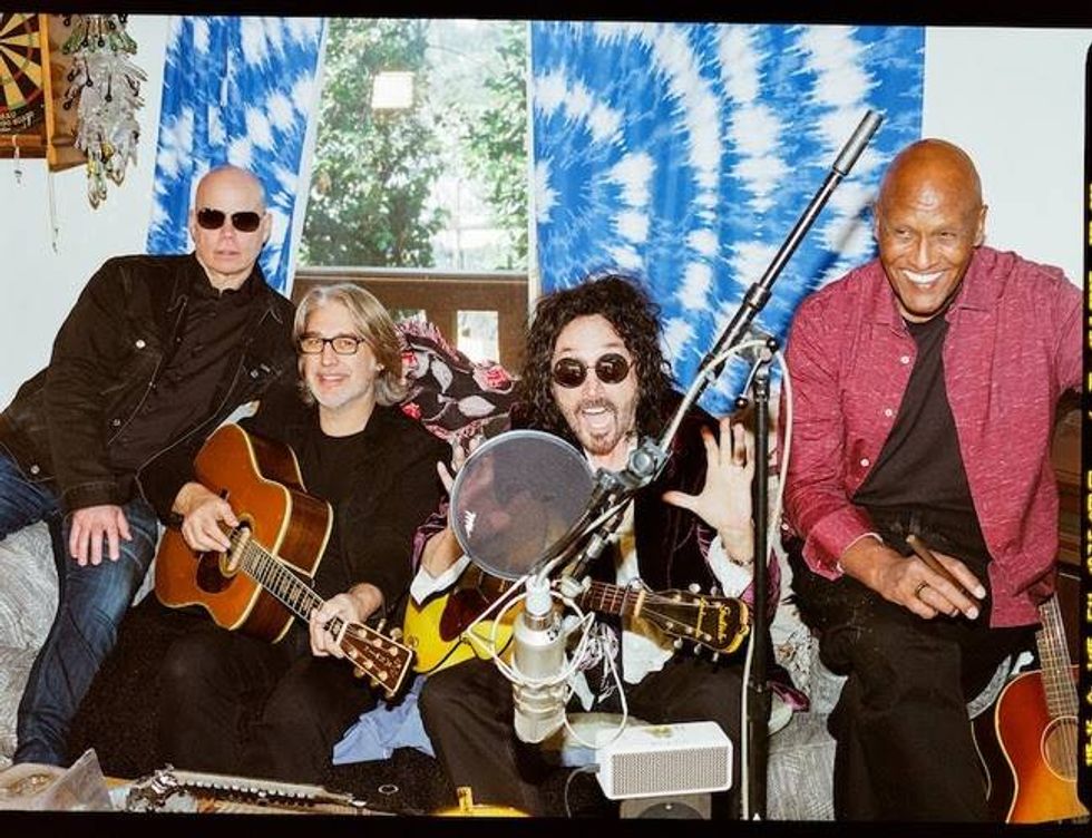 Four musicians sit together in a cozy room, playing guitars and smiling, with colorful decor.